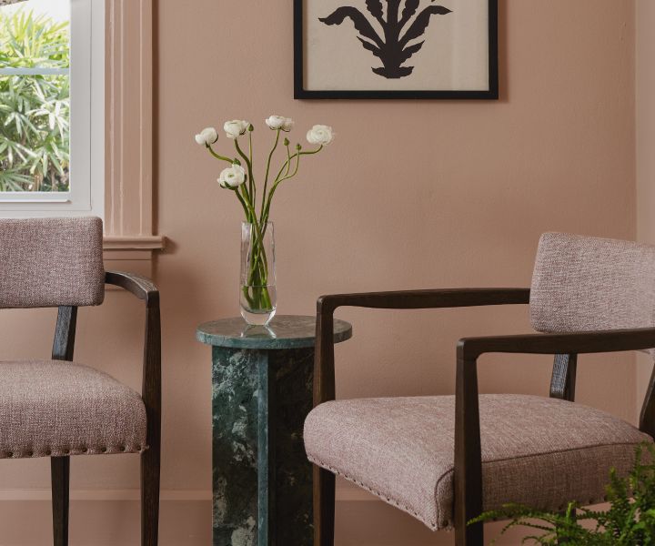 Two upholstered chairs and a marble side table with fresh flowers, against a blush wall